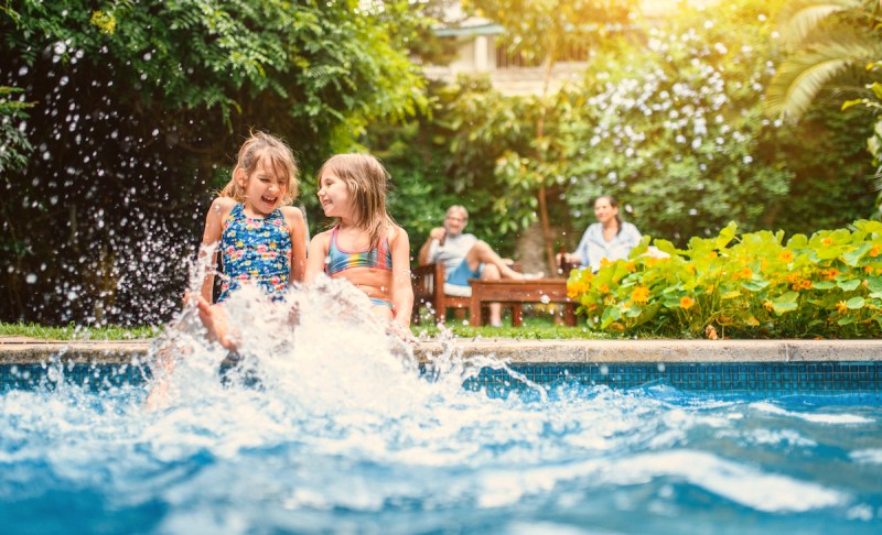 Laughing 6 and 8 year old sisters in swimwear sitting on side of pool and kicking their legs in water.